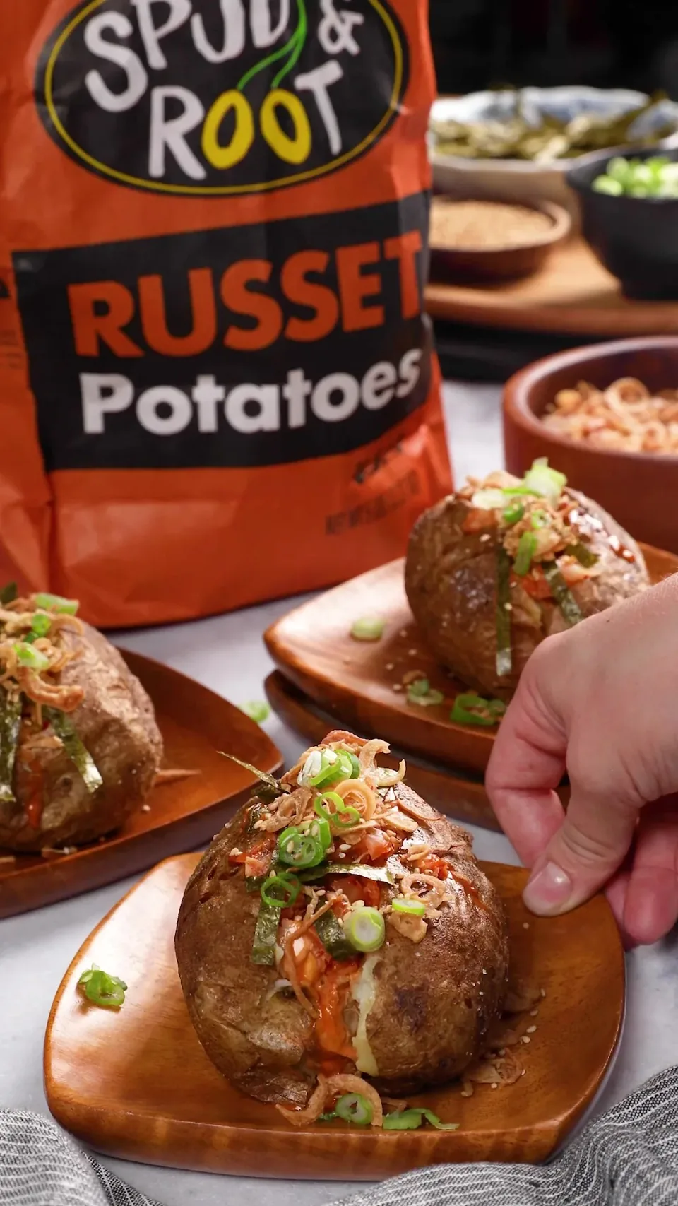 A loaded baked potato on a wooden dish, with more green onion being added, in front of a bag of Spud & Root Russet Potatoes.