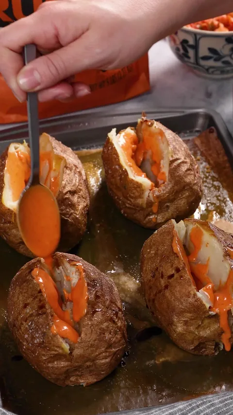 A spoon drizzling orange spicy mayo into the split baked potatoes.