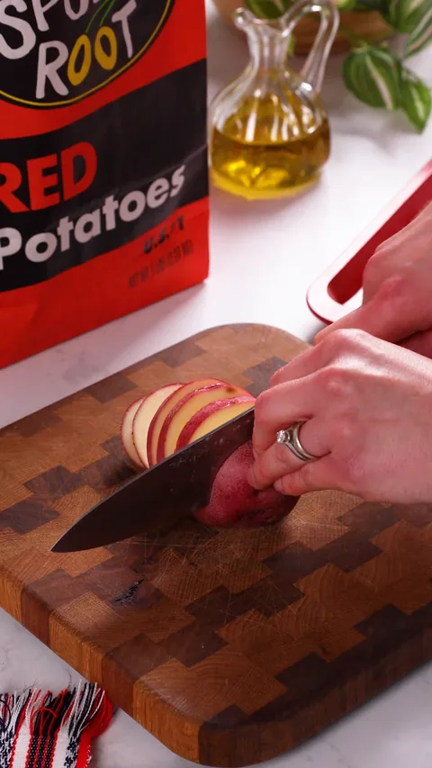 A knife slicing a red potato into thin rounds on a wooden cutting board.