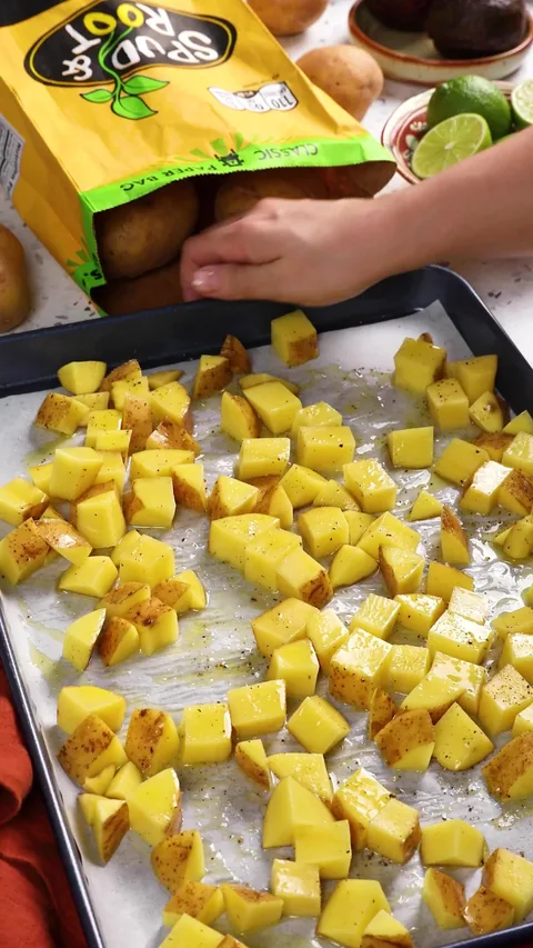 A hand holding the edge of the baking sheet of seasoned potato cubes, spread out in one layer, beside a bag of Spud & Root Gold Potatoes.