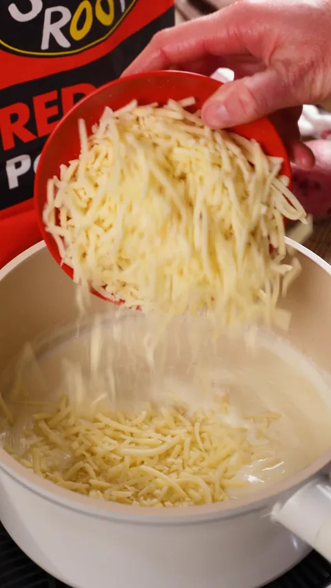 A hand tipping a red bowl of shredded Gruyère into the white sauce.