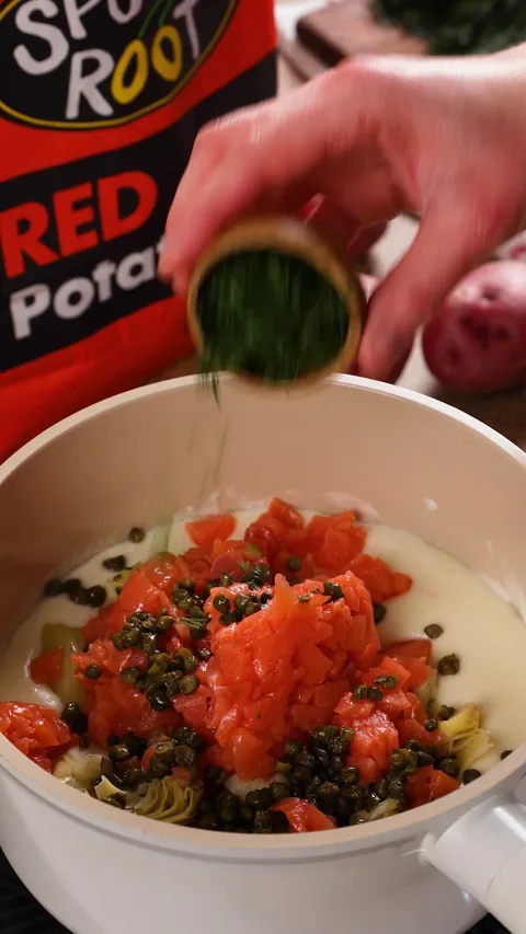 Chopped smoked salmon, artichoke hearts and capers on top of the white sauce, with chopped dill falling from a small bowl.