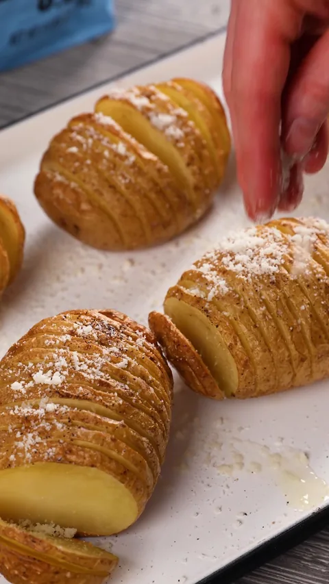 A hand sprinkling grated cheese over golden roasted Hasselback potatoes on a white tray.