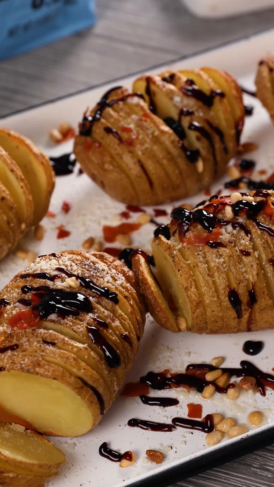 Golden Hasselback potatoes on a white enamel tray, topped with grated cheese, crispy prosciutto and pine nuts and drizzled with balsamic glaze.
