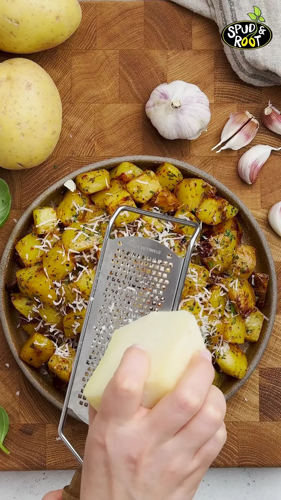 A hand holding a block of Pecorino over a grater above a plate of golden roasted potatoes, seen from above, with grated cheese falling onto them.