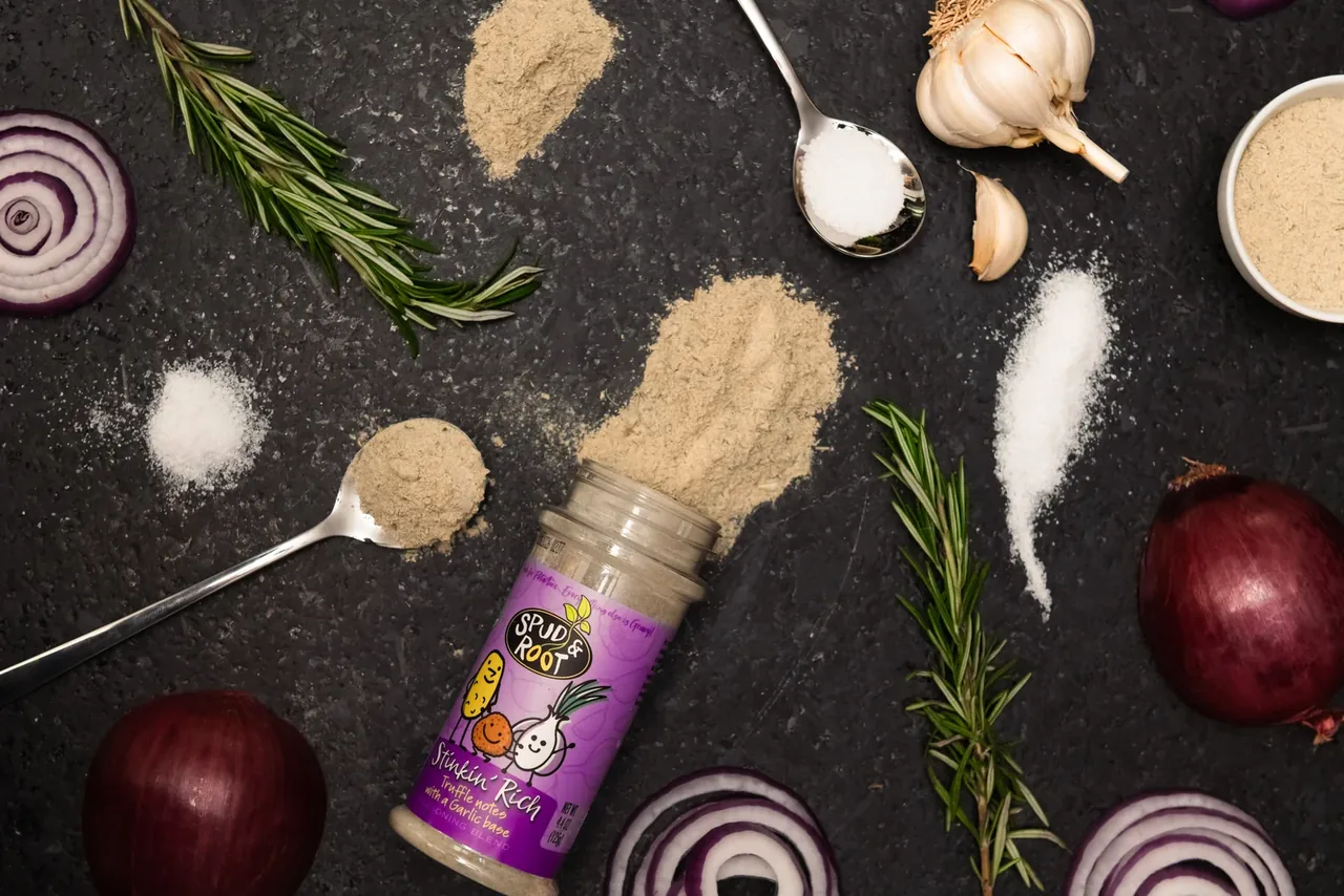 A Stinkin' Rich jar lying on a dark counter, surrounded by garlic, a red onion, rosemary, salt and spoons of seasoning.