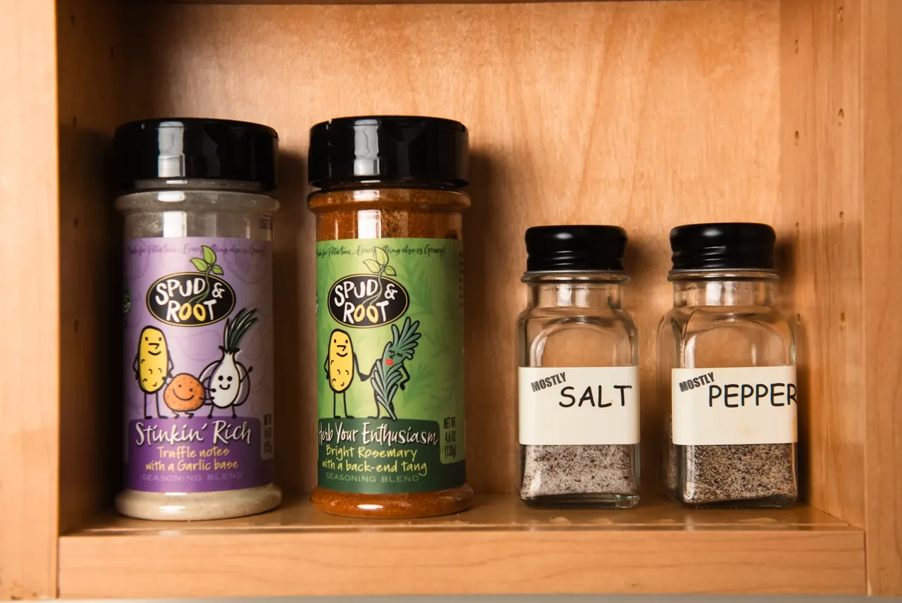 Stinkin' Rich and Herb Your Enthusiasm jars on a kitchen cabinet shelf beside two spice jars labeled Mostly Salt and Mostly Pepper.